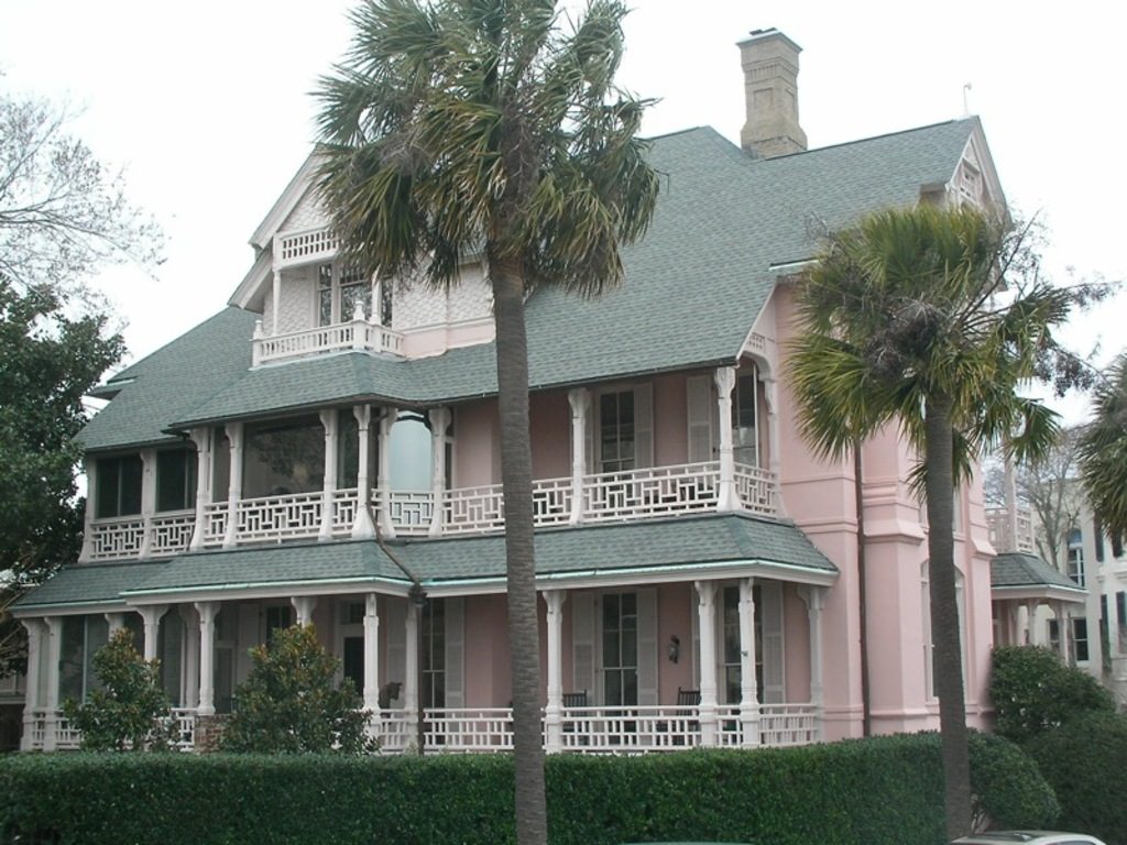 A pink Victorian house with white wraparound porches framed by palm trees