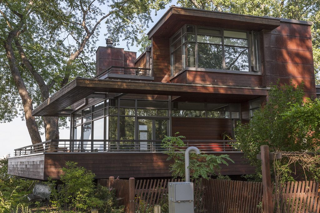 A modern timber-clad house with wide glass windows and a wraparound deck among trees