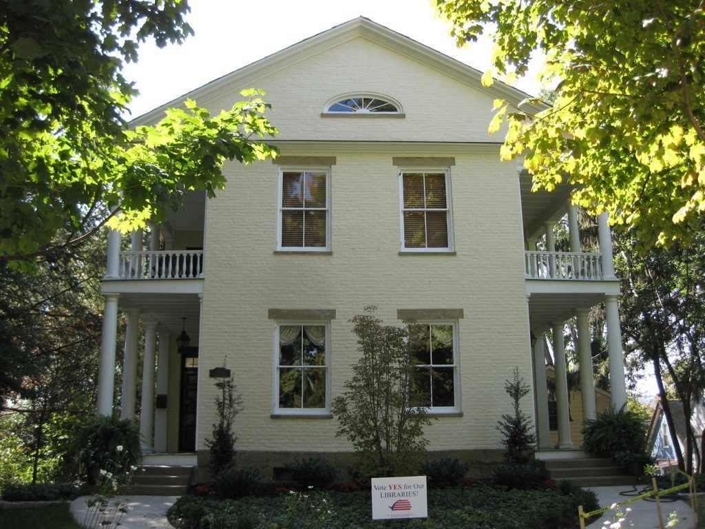 A cream two-storey house with white porches and young trees in the front garden